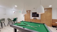 Located at 5580 Thornburn St in Los Angeles, CA, this single-floor community amenity space is thoughtfully designed to offer residents a versatile and stylish environment for relaxation and socializing. The open layout is anchored by light wood-plank flooring and crisp white walls, creating a clean, contemporary aesthetic throughout.

The game area serves as a central highlight, featuring a full-size pool table illuminated by large, dome-shaped pendant lights, alongside a shuffleboard table for additional entertainment. A dedicated media wall with warm wood cabinetry and a wall-mounted flat-screen TV completes this zone.

The wet bar and kitchenette area is well-appointed with white quartz countertops, stainless steel appliances including beverage refrigerators and a microwave, a stainless sink with a modern faucet, and a textured tile backsplash. Open wood shelving with leather-strap hardware adds a stylish, functional touch, while multiple wall-mounted TVs make this a great spot for casual gatherings.

Lounge and seating areas are spread throughout the space, including a comfortable sofa grouping, a row of tan leather bench seating paired with marble-top café tables, and high-top bar tables with eclectic upholstered stools. Lush potted plants and curated wall art add warmth and personality, making this a welcoming destination for the entire community.