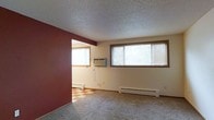 This single-floor space offers a straightforward, functional layout with distinct material transitions between its rooms. The main living area features wall-to-wall carpeting and is finished with a textured popcorn ceiling that adds subtle dimension overhead. Multiple windows fitted with horizontal blinds line the exterior wall, allowing generous natural light to fill the room while offering privacy control. A baseboard heater runs along the base of the exterior wall, providing efficient heating throughout the space. A wall-mounted air conditioning unit ensures year-round climate comfort.

A bold, deep red accent wall frames the open passage to the adjoining room, creating a clear visual separation between the two spaces. This secondary room is finished with a classic checkered vinyl tile floor in a warm tan-and-red pattern, offering a durable, easy-to-clean surface suited for higher-traffic use. The room also benefits from its own window, maintaining consistent natural light throughout. Neutral cream-toned walls across both spaces keep the overall palette simple and versatile, providing a clean backdrop adaptable to a variety of furnishing styles.