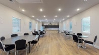 Located at 605 E Fish Creek Rd in Grand Prairie, Texas, this single-story venue offers a thoughtfully designed interior well-suited for gatherings, events, and meetings. The open-concept main hall is generously proportioned, featuring warm wood-look plank flooring, crisp white walls, and a grid of recessed lighting that evenly illuminates the entire space. Multiple windows fitted with white blinds line the walls, allowing natural light to complement the bright overhead lighting and creating a welcoming, airy atmosphere.

The seating area is furnished with round tables and black banquet chairs arranged throughout the hall, providing flexible configurations for a variety of event sizes. High-top tables with bar-height chairs are also present along one side, offering additional casual seating options.

A standout feature of the space is the built-in kitchen area, anchored by a striking peninsula with a stacked-stone base and a white quartz countertop. Dark espresso cabinetry lines the back wall above a tile backsplash, complemented by a stainless steel French-door refrigerator and a utility sink. Decorative accents displayed above the cabinets add a polished, finished look. The kitchen provides excellent functionality for catered events or self-service refreshments. Two entry doors offer convenient access from multiple points.