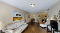 This single-floor residence offers an open-concept layout that integrates the living, dining, and kitchen areas into one cohesive and functional space. The living area is anchored by a comfortable leather sofa and decorated with warm, earth-toned artwork, contributing to a welcoming and relaxed ambiance. Rich, dark hardwood flooring runs throughout, providing a striking visual contrast against the neutral beige walls and white crown molding that frame the space with a clean, classic finish. Ceiling-mounted lighting keeps the interior bright and well-illuminated throughout. The dining area sits just beyond the living space, centered around a classic wooden table and chairs beneath a pendant light, making it ideal for everyday meals or casual gatherings. A decorative wall clock serves as an eye-catching accent in this zone. The kitchen is positioned at the rear of the open layout, featuring warm wood cabinetry, a dark countertop island or breakfast bar, and essential appliances, offering both practicality and style. A hallway visible from the living area suggests access to additional private rooms. Overall, the space balances modern comfort with a warm, inviting aesthetic throughout its single level.
