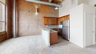 Located at 311 SW Water St in downtown Peoria, Illinois, this one-bedroom, one-bathroom loft showcases a striking industrial aesthetic rooted in its building's historic character. The open-concept layout seamlessly connects the living, dining, and kitchen areas into a single, flowing space defined by soaring ceilings with exposed wood beams and visible ductwork that enhance the loft's urban charm.

The kitchen is a functional centerpiece, featuring rich wood cabinetry, stainless steel appliances including a refrigerator and range, and a peninsula with a neutral countertop that doubles as a breakfast bar. Exposed brick walls frame the kitchen and extend throughout the unit, providing a warm, textural contrast against the crisp white walls and soft carpeted floors.

Large industrial-style windows flood the living area with natural light and offer open views of the surrounding cityscape. A dedicated entrance corridor with a pendant light and original timber elements leads into the main living space, setting the tone for the loft's character.

The bedroom provides a comfortable retreat with a spacious built-in closet and continued carpeted flooring, while two total closets offer ample storage throughout the unit. The bathroom includes a full tub-shower combination. This loft delivers a compelling blend of historic industrial design and modern residential comfort in the heart of Peoria.