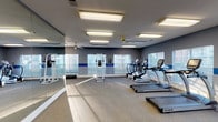This fitness center offers a bright, open workout environment designed to support a well-rounded exercise routine. The space is finished with neutral carpet flooring and gray walls accented by a bold blue horizontal stripe, giving the room a clean, energetic aesthetic. Recessed fluorescent lighting panels are distributed evenly across the ceiling, supplemented by multiple ceiling fans that promote air circulation throughout the space. Large windows line multiple walls, inviting ample natural light and providing views of the surrounding outdoor environment, enhancing the overall atmosphere during workouts. A full-wall mirror along one side of the room amplifies the sense of space and allows users to monitor their form. The cardio equipment section is well-stocked with multiple TRUE-brand treadmills and elliptical cross-trainers, arranged to allow comfortable spacing between machines. Strength training options include a dedicated power tower for bodyweight exercises such as pull-ups and dips. A water fountain is conveniently located near the entry, and a wall-mounted sanitizer dispenser promotes cleanliness. The space also includes a private room accessible via interior doors, adding functional flexibility to the facility. Overall, the fitness center delivers a practical, well-lit, and thoughtfully organized environment for a comprehensive workout experience.