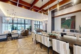 Located at Civic Center Place in Miramar, Florida, this generously sized single-floor clubhouse offers a sophisticated, open-plan amenity space designed with contemporary elegance. The interior is anchored by striking geometric parquet wood flooring that flows seamlessly throughout, complemented by soaring ceilings adorned with rich warm-toned wood beam detailing and recessed lighting with ambient cove illumination.

The main lounge area is furnished with a curated mix of leather and upholstered seating arrangements atop bold patterned area rugs, creating inviting conversational groupings. Dramatic globe and linear pendant chandeliers in both brass and chrome finishes serve as focal points throughout. A large-format flat-screen TV is mounted on a feature wall, providing an entertainment hub within the lounge.

A substantial conference-style dining table with upholstered seating accommodates large gatherings beneath elegant cylindrical pendant lights and exposed wood ceiling beams. Floor-to-ceiling glass curtain walls and expansive windows flood the space with natural light and offer views of the surrounding outdoor areas.

A dedicated billiards room, highlighted by an artistic cluster of molten-glass pendant lights, adds a playful recreational element. White stacked-stone accent walls display curated original artwork throughout, contributing to the space's gallery-like ambiance.