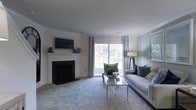 This two-floor home offers a thoughtfully designed living space with a warm, inviting ambiance. The living room serves as a central highlight, featuring a traditional white-mantled fireplace with a sleek, dark surround and a wall-mounted television above — a functional and stylish focal point. Crown molding lines the ceiling, lending a refined architectural touch to the room.

Natural light flows generously through a large sliding glass door framed by elegant blue and white gingham curtains, offering views of a lush green outdoor area. The neutral carpeted flooring and soft grey and cream wall tones create a calming, cohesive palette throughout. A contemporary glass and metallic coffee table anchors the seating area, complemented by a grey sofa accented with blue and textured throw pillows, reinforcing the cool, coordinated color scheme.

A tripod floor lamp beside the seating area adds a modern decorative element. The overall design balances traditional architectural details with contemporary furnishings, resulting in a comfortable and visually polished living environment across both floors.