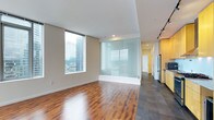 This contemporary single-floor residence showcases a clean, modern design with an open-concept layout that maximizes both space and functionality. The living area is anchored by rich hardwood flooring that flows seamlessly throughout, while a striking frosted glass partition adds a stylish architectural element that defines spaces without sacrificing light or openness. Floor-to-ceiling windows flood the interior with natural light and frame sweeping urban cityscape views, creating a dramatic connection to the surrounding skyline. The transition from the living area into the kitchen zone is marked by a shift to sleek dark tile flooring, complementing the warm yellow cabinetry and stainless steel appliances that define the culinary space. Track lighting overhead provides functional illumination throughout the kitchen corridor. Neutral wall tones and white ceilings keep the interiors feeling bright and airy, while the minimalist aesthetic ensures the architectural details remain the focal point. A hallway extends toward additional rooms, maintaining the clean lines and modern finishes consistent throughout the residence. The overall ambiance is sophisticated, light-filled, and distinctly urban.