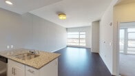 This contemporary single-floor residence offers a thoughtfully designed open-concept layout that seamlessly connects the living, dining, and kitchen areas. Rich dark hardwood flooring runs throughout the main living spaces, providing a striking contrast to the crisp white walls and ceilings.

The kitchen is a standout feature, outfitted with a full suite of stainless steel appliances — including a refrigerator, range, and microwave — complemented by white cabinetry, granite countertops, and a peninsula with an integrated sink. Recessed lighting illuminates the space, adding warmth and functionality.

The open living area is bathed in natural light through large floor-to-ceiling windows that frame expansive city views, enhancing the sense of openness and scale. A bedroom features plush carpeting and direct access to a private balcony through wide sliding glass doors, offering an elevated outdoor vantage point with panoramic urban vistas.

The bathroom is finished with neutral tile flooring, a granite-topped vanity with white cabinetry, and a well-lit mirror. The consistent use of granite surfaces and stainless steel hardware throughout lends a cohesive, modern aesthetic. Clean lines, neutral tones, and an abundance of natural light define the overall ambiance of this well-appointed urban residence.