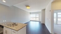 This contemporary single-floor residence offers a thoughtfully designed open-concept layout that seamlessly connects the living, dining, and kitchen areas. Rich dark hardwood flooring runs throughout the main living spaces, providing a striking contrast to the crisp white walls and ceilings.

The kitchen is a standout feature, outfitted with a full suite of stainless steel appliances — including a refrigerator, range, and microwave — complemented by white cabinetry, granite countertops, and a peninsula with an integrated sink. Recessed lighting illuminates the space, adding warmth and functionality.

The open living area is bathed in natural light through large floor-to-ceiling windows that frame expansive city views, enhancing the sense of openness and scale. A bedroom features plush carpeting and direct access to a private balcony through wide sliding glass doors, offering an elevated outdoor vantage point with panoramic urban vistas.

The bathroom is finished with neutral tile flooring, a granite-topped vanity with white cabinetry, and a well-lit mirror. The consistent use of granite surfaces and stainless steel hardware throughout lends a cohesive, modern aesthetic. Clean lines, neutral tones, and an abundance of natural light define the overall ambiance of this well-appointed urban residence.