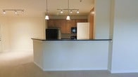 This single-floor residence offers a thoughtfully designed open-concept layout that promotes a seamless flow between the kitchen and living areas. The kitchen is a standout feature, anchored by a raised breakfast bar with a dark granite countertop that provides both a casual dining space and a natural visual divider between the cooking and living zones. Warm honey-toned wood cabinetry lines the kitchen walls, offering generous storage, while the space is equipped with full-sized appliances, including a refrigerator, built-in microwave, and dishwasher. Overhead, a combination of pendant lights and a track lighting fixture illuminates the kitchen with a warm, inviting glow. The living area is finished with plush wall-to-wall carpeting that adds comfort underfoot, while neutral-toned walls create a clean and versatile backdrop throughout the space. The open sightlines between rooms enhance the overall sense of space, making the layout feel bright and connected. The single-floor plan ensures easy navigation and accessibility throughout the residence, making it a practical and comfortable living environment suitable for a variety of lifestyles.
