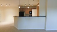 This single-floor residence offers a thoughtfully designed open-concept layout that promotes a seamless flow between the kitchen and living areas. The kitchen is a standout feature, anchored by a raised breakfast bar with a dark granite countertop that provides both a casual dining space and a natural visual divider between the cooking and living zones. Warm honey-toned wood cabinetry lines the kitchen walls, offering generous storage, while the space is equipped with full-sized appliances, including a refrigerator, built-in microwave, and dishwasher. Overhead, a combination of pendant lights and a track lighting fixture illuminates the kitchen with a warm, inviting glow. The living area is finished with plush wall-to-wall carpeting that adds comfort underfoot, while neutral-toned walls create a clean and versatile backdrop throughout the space. The open sightlines between rooms enhance the overall sense of space, making the layout feel bright and connected. The single-floor plan ensures easy navigation and accessibility throughout the residence, making it a practical and comfortable living environment suitable for a variety of lifestyles.