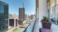 This expansive balcony is a standout feature of the space, positioned at a significant elevation above the Chicago cityscape and offering unobstructed panoramic views of some of the city's most iconic landmarks. The Chicago River winds directly below, framed by the distinctive corncob towers of Marina City and the soaring glass facade of Trump International Hotel & Tower dominating the skyline. The downtown Chicago skyline stretches broadly across the horizon under open skies, creating a dramatic urban backdrop.

The balcony itself is designed with clean, modern detailing. A continuous glass railing system runs the full length of the outdoor space, preserving sightlines while maintaining a sense of openness and safety. The curved white exterior wall of the building adds an architectural softness to the space. Large rectangular planter boxes positioned along the railing introduce greenery and seasonal flowers, bringing a natural element that contrasts beautifully with the steel-and-glass cityscape. Seating is available, making this an ideal space for relaxing or entertaining while taking in the remarkable views. The overall ambiance is polished, cosmopolitan, and visually striking.