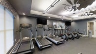 This single-level fitness space is designed to provide a functional and comfortable workout environment. The room is outfitted with a row of treadmills and elliptical machines from Life Fitness, offering users a variety of cardio options. The equipment is arranged along one wall, maximizing the use of available floor space while maintaining an open, uncluttered feel.

The interior is finished with dark gray walls that lend a modern, focused atmosphere to the space. Rubber flooring runs throughout, providing both durability and traction suitable for an active environment. Overhead, a combination of recessed lighting and linear LED fixtures ensures even, bright illumination across the room. A large ceiling fan adds to the ventilation and comfort of the space during workouts.

Mounted flat-screen televisions are positioned at convenient viewing angles along the walls, allowing users to stay entertained during their sessions. A window provides a degree of natural light, while the overall layout and design reflect a clean, contemporary aesthetic well-suited to a dedicated fitness facility.