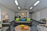 Located at 6655 Mount Zion Blvd in Morrow, Georgia, this single-floor community space offers a welcoming and well-appointed environment designed for comfortable gatherings. The main area is defined by soaring vaulted ceilings fitted with multiple skylights and recessed panel lighting, flooding the room with abundant natural and artificial light. Ceiling fans provide additional comfort throughout the space.

The open layout is thoughtfully arranged to serve multiple functions. A generously sized lounge area features a plush sofa, accent armchairs with colorful throw pillows, and a round wooden coffee table, creating an inviting setting for relaxed conversation. Adjacent to this is a formal dining area with a dark wood round table and elegantly upholstered chairs, suitable for meetings or shared meals. A classic console table along the wall adds a refined decorative touch.

Underfoot, a bold patterned carpet in grey and teal tones adds a vibrant, modern aesthetic that ties the space together. White wainscoting runs along the lower walls, complementing the neutral, light-toned upper walls and contributing to a clean, polished appearance. Large windows offer views of surrounding greenery, enhancing the airy ambiance.

A compact kitchenette off the main area includes white cabinetry, speckled countertops, and a double stainless steel sink with a gold-finish faucet, providing practical support for event catering and everyday use.