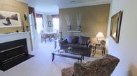 This single-floor space showcases a seamlessly connected living and dining area with a warm, traditional aesthetic. The living area is anchored by a plush, striped sofa in rich tones of blue and burgundy, complemented by a floral-upholstered armchair and a sleek glass-topped coffee table with wrought iron detailing. A matching side table holds a classic table lamp, adding a soft, inviting glow to the space. The walls are finished in a warm taupe tone, accented by white crown molding that runs throughout, lending an air of refinement. Decorative shelving displays elegant vases with sculptural dried branch arrangements, adding height and visual interest to the walls. The dining area, visible through a wide opening, features a traditional wooden table surrounded by Queen Anne-style chairs, framed by windows dressed in rich, patterned valances that allow generous natural light to fill the room. Light-toned wall-to-wall carpeting runs throughout both spaces, contributing to the overall sense of warmth and comfort. The open layout and cohesive design elements create a welcoming, classically styled living environment.
