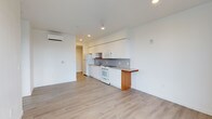 This single-floor space showcases a bright, open-concept design with a minimalist aesthetic. The layout seamlessly integrates the living area and kitchen, creating a spacious and functional environment. Light-toned hardwood flooring flows throughout the unit, complementing the crisp white walls and ceiling that amplify the sense of openness.

The kitchen is designed for practicality and efficiency, featuring white cabinetry extending to the ceiling for ample storage, a grey tile backsplash, and white appliances including a refrigerator, oven, microwave, and dishwasher. A compact kitchen bar with a warm wood-toned countertop provides additional workspace and casual dining potential. Recessed lighting illuminates the kitchen area, while a wall-mounted mini-split HVAC unit ensures year-round climate comfort.

The open living area offers a generous, flexible footprint with clean lines and neutral tones, making it easily adaptable to a variety of furniture arrangements. A hallway at the rear of the space leads to additional rooms, suggesting a well-organized floor plan. Overall, the space presents a fresh, modern atmosphere ideal for comfortable, contemporary living.