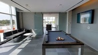 This single-floor recreational space is designed with a clean, contemporary aesthetic. The focal point of the room is a full-sized pool table with a slate-blue felt surface and dark wood frame, centrally positioned on large-format grey ceramic tile flooring. The billiards area is complemented by a casual lounge corner featuring dark upholstered seating and a sleek round white side table, ideal for socializing between games.

The walls are clad in textured stone-effect panels in muted grey tones, contributing to the space's modern and polished character. Recessed ceiling lighting provides even illumination throughout, maintaining a bright and inviting atmosphere.

A standout architectural feature is the pair of sliding glass barn doors that divide the interior from a corridor leading to an outdoor terrace. Large windows beyond the doorway flood the transitional area with natural light and offer views of the exterior, creating a seamless indoor-outdoor connection. The terrace, visible through the glazing, appears to include additional furnishings, extending the usable recreational area outdoors.