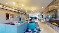 Luxor is a thoughtfully designed, single-floor amenity space that seamlessly blends social, recreational, and lounge environments into one dynamic destination. The welcoming lobby greets visitors with a bold, contemporary aesthetic — featuring sculptural brass-and-glass cluster chandeliers, vibrant teal and blue sectional seating, tufted ottomans, and a striking striped area rug over polished concrete floors. Decorative laser-cut metal panels frame the space, lending an artistic, modern edge.

At the heart of the facility is the Luxor bar and lounge area, anchored by a curved, dark-stone bar with wraparound seating, a coffered ceiling detail overhead, and warm wood accent walls. Nearby, the "Pour" bar station and surrounding café-style tables create an inviting gathering hub bathed in natural light from floor-to-ceiling windows.

A dedicated game room delivers entertainment with classic arcade machines, a foosball table, a pool table, and a witty hand-lettered wall mural. A glowing neon "Game On" sign sets the tone, while a stainless steel counter bar adds a functional hangout spot.

A quieter lounge nook features a sleek linear fireplace with a stone surround, large-scale artwork, and a curated arrangement of lounge chairs — ideal for relaxed conversation or unwinding in style.