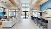 This thoughtfully designed single-floor communal space blends contemporary and rustic elements to create a welcoming, upscale lounge environment. The space is immediately defined by its dramatic double-height ceilings, adorned with large, exposed wood beams that add warmth and architectural character against crisp white walls.

Expansive floor-to-ceiling black-framed windows and glass doors flood the interior with natural light while offering unobstructed views of the outdoor pool and courtyard. Large-format light tile flooring runs throughout, enhancing the open, airy feel of the space.

The living area centers around a striking full-height tiled feature wall, which houses a linear gas fireplace and a mounted flat-screen television. A teal accent wall with floating wood shelves and built-in cabinetry adds a bold pop of color, while a sputnik-style chandelier contributes a modern touch. Comfortable seating arrangements, including neutral sofas and accent chairs, invite relaxed gathering.

The kitchen area features a generous bar-height island with seating for several, pendant lighting with gold-toned interiors, a stainless steel refrigerator, subway tile backsplash, and dark wood cabinetry. Transitional seating nooks near the glass walls provide additional areas for casual conversation or quiet retreat, all with views of the outdoor amenities beyond.