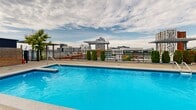 Located at 650 Wellington in Montréal's dynamic Griffintown neighbourhood, this rooftop pool terrace is a sophisticated outdoor amenity space designed for relaxation and leisure. The centrepiece is a generously sized outdoor swimming pool with vivid blue tile finishes, complemented by a dedicated wading or spa pool nearby. Surrounding the pool, an expansive sundeck is lined with rows of loungers, providing ample space for sunbathing beneath open skies. Structural pergolas punctuate the terrace, offering shaded retreats and architectural interest, while natural wood privacy screens add warmth and definition to the space. Lush, tiered planter beds filled with a variety of greenery and ornamental plants are integrated throughout, softening the hardscape and creating a resort-like atmosphere. An enclosed glass pavilion structure at one end of the terrace extends the usable space and provides shelter from the elements. Glass and metal railings frame the perimeter, ensuring unobstructed panoramic views of the Montréal skyline, including the downtown core, surrounding neighbourhoods, and historic architecture. The open-sky setting allows abundant natural light throughout the day, making this rooftop terrace a standout urban retreat that seamlessly blends modern design with vibrant city living.