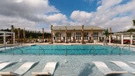 Located in Summerville, South Carolina, this single-story outdoor amenity facility offers a resort-inspired pool experience designed for relaxation and recreation. The centerpiece is a generously sized swimming pool with crystal-clear blue water, flanked by in-water sun shelves ideal for lounging. Rows of towering palm trees line the pool deck, lending a coastal ambiance to the space and providing natural shade throughout the day. The surrounding pool deck is finished in light-colored pavers, offering ample room for chaise lounges and seating areas. A pergola structure on one side of the deck provides additional shade, while multiple lounge chairs are thoughtfully arranged to take full advantage of the open sky and natural light. The adjacent clubhouse building features classic white board-and-batten siding topped with a traditional pitched roof, and is fronted by a wall of large, grid-paned French doors that seamlessly connect the interior to the pool deck. These expansive glass doors invite abundant natural light and maintain a visual connection between indoor and outdoor spaces. The lush treeline of South Carolina pines forms a natural backdrop, enhancing the sense of privacy and tranquility throughout this beautifully landscaped amenity area.