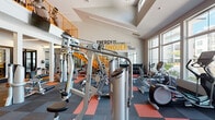 This two-story fitness center is thoughtfully designed to accommodate a wide range of workout styles and fitness goals. The main floor is expansive and open, featuring soaring double-height ceilings that create an energizing atmosphere. Large floor-to-ceiling windows along the perimeter flood the space with natural light while offering views of the surrounding community. The flooring consists of a bold patchwork of charcoal and red modular carpet tiles, adding visual energy to the workout environment.

The main floor is densely equipped with a comprehensive selection of strength machines, free weights, cable systems, rowing machines, and ellipticals. A dedicated dumbbell rack with color-coded weights lines one wall alongside medicine balls and kettlebells. Bold motivational typography — including phrases like "Energy and Persistence Conquer All Things" and "Ride It Like You Stole It" — is applied directly to the walls, reinforcing an active and determined atmosphere.

A separate studio room, accessible through sleek black-framed glass doors, features hardwood-style flooring, a full-length mirror wall, cycling bikes, and a mounted television, making it ideal for group or solo classes.

A striking orange staircase leads to the second floor, where additional cardio equipment, including rowing machines and upright bikes, is arranged along an open mezzanine with bright linear lighting overhead.