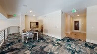 Located at 7770 Farr St in Charleston, SC, this single-floor amenity lounge is designed to foster social activity and relaxed recreation. The space is unified throughout by a striking, boldly patterned carpet in deep teal, navy, and neutral tones that sets a lively yet sophisticated atmosphere against the clean white walls and light-colored ceilings.

The centerpiece of the main area is a full-size billiards table, illuminated overhead by an industrial-style pendant light fixture that adds a warm, focused glow. Adjacent to it, a shuffleboard table extends the game room experience, offering additional entertainment options for residents or guests. Pendant lighting continues across the space, complementing the recessed ceiling lights that provide even, ambient illumination throughout.

Toward one end, a dedicated lounge seating area features patterned accent chairs and a central coffee table, accented by a coastal landscape painting on the wall — a nod to the surrounding Lowcountry environment. At the opposite end, a casual game corner includes a dining-height table with upholstered chairs and a wall-mounted board game display, creating an inviting spot for tabletop activities.

Large windows along one wall allow natural light to filter in, adding brightness and openness to the interior. The overall design balances functionality with a refined, contemporary aesthetic suited to a welcoming community gathering space.