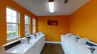 This communal laundry facility is designed with both functionality and energy in mind. The room immediately draws attention with its bold, warm orange walls, which lend a lively and welcoming atmosphere to an otherwise utilitarian space. A decorative floral canvas print adds a touch of personality to the room, complementing the vibrant color scheme.

The facility is lined on both sides with rows of white top-load washing machines and dryers, providing ample capacity for multiple users. The machines are neatly arranged along the walls, maximizing the use of available floor space while maintaining clear, open access through the center of the room. The dark tile flooring offers a practical, easy-to-clean surface that contrasts cleanly with the white appliances.

Natural light filters into the room through two double-hung windows on one wall, brightening the space during daytime hours. Overhead, a fluorescent ceiling fixture ensures consistent, even illumination throughout. A ceiling-mounted HVAC vent contributes to comfortable air circulation within the space. The textured ceiling and clean white trim provide a finished look that completes the room's practical yet cheerful design. Overall, the facility is well-maintained and thoughtfully arranged to serve the needs of its users efficiently.