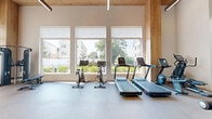 This fitness center on East Ave in Norwalk, Connecticut, offers a thoughtfully designed exercise space that combines functionality with modern aesthetics. The room is defined by soaring double-height ceilings accented with warm wood paneling, creating an open and energizing atmosphere. Large windows along one wall flood the space with natural light while framing views of the surrounding neighborhood greenery, making workouts feel connected to the outdoors.

The exercise room is outfitted with a comprehensive range of equipment to support both cardio and strength training. Cardio options include multiple treadmills and stationary cycling bikes, each equipped with digital display screens. The strength training area features a cable machine, a Smith machine with a full weight rack, a multi-function cable system by TRUE Fitness, and adjustable benches. Full-length mirrors along one wall enhance the sense of space and assist with form during workouts.

Durable rubberized flooring covers the entire area, providing safety and comfort underfoot. Black-framed interior glass partitions add an industrial-modern architectural detail, complementing the clean white walls and wood ceiling elements. Pendant lighting provides additional illumination alongside recessed ceiling fixtures. The layout offers ample open floor space between equipment stations, ensuring comfortable movement throughout the room.