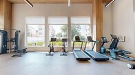 This fitness center on East Ave in Norwalk, Connecticut, offers a thoughtfully designed exercise space that combines functionality with modern aesthetics. The room is defined by soaring double-height ceilings accented with warm wood paneling, creating an open and energizing atmosphere. Large windows along one wall flood the space with natural light while framing views of the surrounding neighborhood greenery, making workouts feel connected to the outdoors.

The exercise room is outfitted with a comprehensive range of equipment to support both cardio and strength training. Cardio options include multiple treadmills and stationary cycling bikes, each equipped with digital display screens. The strength training area features a cable machine, a Smith machine with a full weight rack, a multi-function cable system by TRUE Fitness, and adjustable benches. Full-length mirrors along one wall enhance the sense of space and assist with form during workouts.

Durable rubberized flooring covers the entire area, providing safety and comfort underfoot. Black-framed interior glass partitions add an industrial-modern architectural detail, complementing the clean white walls and wood ceiling elements. Pendant lighting provides additional illumination alongside recessed ceiling fixtures. The layout offers ample open floor space between equipment stations, ensuring comfortable movement throughout the room.