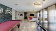 This single-floor community amenity facility in Carbondale, Illinois — Home of the Salukis — offers a diverse range of thoughtfully designed spaces for recreation, productivity, and socializing. The entrance lobby sets an energetic tone with light wood flooring and a striking feature wall adorned with mounted bicycles against a dark geometric mural. The open game and lounge area serves as the social heart of the facility, combining a red felt pool table, ping-pong table, and a stylish lounge seating area. The lounge features bold fuchsia velvet accent chairs, a grey sofa with red throw pillows, a sleek circular pendant light fixture, a wood-panel accent wall with a wall-mounted flat-screen TV, and local Carbondale artwork. A café-style bar area with black subway tile, pendant lighting, bar stools, and a coffee station provides a welcoming refreshment spot. The computer lab offers workstations along built-in desks beneath an arched window that brings in natural light. A spacious study and meeting room with vibrant orange and teal accent walls, modular tables, and ample seating supports collaborative work. Completing the amenities is a private screening room outfitted with tiered, stadium-style recliner seating, sconce lighting, and classic movie poster décor for an immersive cinema experience.