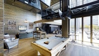 This single-floor Dallas entertainment space blends industrial and modern design elements to create a vibrant, multi-functional gathering environment. The interior is anchored by a wood-framed billiards table at the center, positioned beneath a striking array of large mounted flat-screen televisions suspended from the soaring double-height ceiling. Exposed brick walls add texture and warmth, contrasting beautifully with the sleek black metal railings of an open mezzanine-style overlook above. Light hardwood flooring runs throughout, enhancing the bright, open atmosphere. A lounge area with comfortable gray seating and an antler chandelier adds a rustic touch to the otherwise industrial aesthetic. High-top bar seating and casual tables provide additional gathering spots, while floor-to-ceiling windows along one side flood the space with abundant natural light and offer views of the outdoors. A staircase with black metal railings leads to the upper overlook level, adding vertical visual interest. The overall ambiance is energetic yet inviting, making it well-suited for social events, casual recreation, and communal gatherings.
