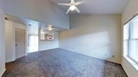 This two-floor residence offers a thoughtfully designed layout that balances open living spaces with functional flow. The main living area immediately impresses with its vaulted ceilings, which lend a sense of spaciousness and airiness to the room. A ceiling fan is centrally mounted, providing both comfort and a subtle focal point overhead. The room is finished with wall-to-wall carpeting in a neutral tone, creating a warm and inviting atmosphere throughout.

A pass-through window connects the living area to the kitchen, allowing natural sightlines between the two spaces and making the floor plan feel open and communicative. The kitchen area, visible from the living room, features cabinetry and overhead lighting, providing a practical and well-lit workspace. A window in the adjoining area admits natural light, brightening the interior and enhancing the overall sense of openness.

The neutral color palette carried throughout the main level — from the beige walls to the toned carpeting — creates a clean, versatile backdrop suitable for a variety of interior styles. With two floors of living space, this home provides ample room for both everyday living and private retreats, making it a well-rounded and functional residence.