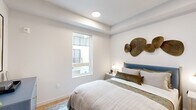 This thoughtfully designed single-floor space offers a cohesive blend of modern aesthetics and functional living. The bedroom serves as the centerpiece, featuring warm light wood flooring, crisp white walls, and a tray ceiling with recessed lighting that adds subtle architectural depth. A large window allows natural light to filter in while offering views of the surrounding exterior. The room includes a built-in closet with a sliding door, providing practical storage, and is complemented by clean wall space that lends itself well to décor. Adjacent to the bedroom is a sleek, modern bathroom finished in a bright, minimalist style. It features warm wood-tone flooring that carries through from the bedroom, a floating vanity with a white countertop and undermount sink, an oval wall mirror with a slim black frame, and wall sconce lighting. The layout is efficient yet comfortable, with ample wall space accented by framed artwork. Together, the rooms reflect a consistent design language — neutral palettes, quality materials, and clean architectural lines — creating a calm, contemporary ambiance throughout the space.