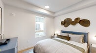 This thoughtfully designed single-floor space offers a cohesive blend of modern aesthetics and functional living. The bedroom serves as the centerpiece, featuring warm light wood flooring, crisp white walls, and a tray ceiling with recessed lighting that adds subtle architectural depth. A large window allows natural light to filter in while offering views of the surrounding exterior. The room includes a built-in closet with a sliding door, providing practical storage, and is complemented by clean wall space that lends itself well to décor. Adjacent to the bedroom is a sleek, modern bathroom finished in a bright, minimalist style. It features warm wood-tone flooring that carries through from the bedroom, a floating vanity with a white countertop and undermount sink, an oval wall mirror with a slim black frame, and wall sconce lighting. The layout is efficient yet comfortable, with ample wall space accented by framed artwork. Together, the rooms reflect a consistent design language — neutral palettes, quality materials, and clean architectural lines — creating a calm, contemporary ambiance throughout the space.