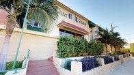 Casa Grande is a Spanish-influenced multi-unit building identified by its warm yellow stucco exterior, brick accents, red tile roofline, and teal green architectural detailing that lends the property a distinctive, cohesive character. The building's façade is framed by mature palm trees, manicured hedges, and colorful plantings, creating an inviting street presence.

The unit featured here is situated on a single floor and includes access to a private outdoor balcony or walkway corridor. This outdoor area is finished with a pink-toned concrete floor and enclosed by decorative teal metalwork—a combination of expanded mesh panels and ornamental circular lattice gate detailing—providing both privacy and visual interest. A classic hanging lantern-style light fixture adds a warm, traditional touch to the corridor.

From the balcony, residents enjoy open views of the surrounding greenery, including towering palms and dense shrub landscaping that offer a natural sense of seclusion. The teal railings and metalwork gates continue the building's signature color palette throughout the exterior spaces, ensuring a unified aesthetic. The overall ambiance blends Southern California outdoor living with enduring Spanish Colonial Revival design elements.