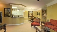 This single-floor space showcases an open-plan layout that seamlessly connects the kitchen, dining area, and living room, creating a cohesive and inviting atmosphere. The kitchen features white cabinetry, a dark countertop breakfast bar with a curved edge, and recessed lighting beneath a tray ceiling detail. Pendant lights hang above the bar, providing both functional task lighting and a decorative touch. White appliances and upper cabinets with decorative accents complete the kitchen's classic aesthetic.

The dining area sits adjacent to the kitchen, centered around a dark wood table and chairs, accented by a chandelier overhead and warm yellow walls. A large framed painting adds a focal point to the space, while wall-mounted decorative plates contribute to the traditional character of the room. White chair rail molding lines the walls, adding architectural detail throughout.

The living area flows naturally from the dining space, furnished with an ornate red upholstered armchair and a glass-topped side table, reflecting a traditional interior style. Soft, neutral carpeting runs throughout, tying the spaces together and contributing to the overall warm, comfortable ambiance of the home.