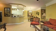 This single-floor space showcases an open-plan layout that seamlessly connects the kitchen, dining area, and living room, creating a cohesive and inviting atmosphere. The kitchen features white cabinetry, a dark countertop breakfast bar with a curved edge, and recessed lighting beneath a tray ceiling detail. Pendant lights hang above the bar, providing both functional task lighting and a decorative touch. White appliances and upper cabinets with decorative accents complete the kitchen's classic aesthetic.

The dining area sits adjacent to the kitchen, centered around a dark wood table and chairs, accented by a chandelier overhead and warm yellow walls. A large framed painting adds a focal point to the space, while wall-mounted decorative plates contribute to the traditional character of the room. White chair rail molding lines the walls, adding architectural detail throughout.

The living area flows naturally from the dining space, furnished with an ornate red upholstered armchair and a glass-topped side table, reflecting a traditional interior style. Soft, neutral carpeting runs throughout, tying the spaces together and contributing to the overall warm, comfortable ambiance of the home.