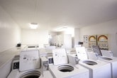 This shared laundry facility is a single-floor, self-contained utility space designed to serve multiple users efficiently. The room is finished with white walls and light tile flooring, giving it a clean and functional aesthetic throughout. Overhead fluorescent lighting ensures the space is well lit at all hours.

The facility is outfitted with a bank of top-loading washing machines arranged in a row, providing ample capacity for simultaneous use. On the opposite side of the room, stacked dryer units offer a space-efficient solution for drying. Coin-operated laundry vending dispensers are mounted on the wall, offering detergent and other laundry essentials for added convenience. Instructional signage for both washing and drying is posted prominently on the walls.

A built-in marble-style counter runs along one wall beneath a large window, offering a dedicated surface for folding and sorting laundry. Natural light filters in through this window, providing a pleasant view of the exterior. A secondary glass-panel door offers an additional point of entry. Seating is available for those waiting, and a security camera is installed on the ceiling, ensuring a safe environment. The space is practical, well-organized, and designed to meet the everyday laundry needs of its users.