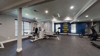 This fitness center is a thoughtfully designed single-floor space built to support a complete workout experience. The layout is open and spacious, with durable rubberized flooring throughout that provides both safety and comfort underfoot during exercise. Recessed lighting illuminates the entire area evenly, while multiple windows fitted with blinds allow natural light to filter in, keeping the environment bright and energizing.

The main workout area is well-stocked with a multi-station cable and weight machine, flat benches, a stationary bike, a treadmill, an elliptical trainer, and an assortment of dumbbells and medicine balls neatly arranged along the wall. A large floor-to-ceiling mirror spans one wall, enhancing the sense of space and allowing users to monitor their form during exercise. Structural columns with clean white detailing are integrated seamlessly into the layout.

A bold navy accent wall serves as a focal point, displaying motivational typography in yellow — featuring words such as "Believe," "Discipline," "Strong," "Consistency," and "Secret" — adding an inspiring atmosphere to the space. A vibrant abstract mural decorates the entryway wall, creating a visually dynamic transition into the fitness area. The entry zone includes a bench and utility station near a glass-paneled door. Crown molding and textured ceilings add a polished architectural finish throughout.