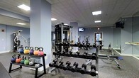 This fitness center is a thoughtfully designed workout facility built to accommodate a wide range of exercise needs. The space features a dropped ceiling with recessed panel lighting that provides bright, even illumination throughout. The walls are painted in a cool, neutral blue-grey tone, contributing to a focused and energizing atmosphere, while rubber flooring offers both durability and safety underfoot.

The strength training area is well-stocked with a full rack of dumbbells spanning a broad range of weights, complemented by kettlebells and medicine balls neatly organized on dedicated storage racks. A multi-functional cable machine serves as a centerpiece for resistance training, with large mirrors mounted along the wall to assist with form and to visually expand the space.

The cardio section is equipped with multiple machines, including ellipticals and upright exercise bikes, positioned to allow comfortable spacing between users. A wall-mounted flat-screen television is available for entertainment during workouts.

Natural light filters in through a window on the far wall, adding a welcoming element to the otherwise interior-focused layout. An emergency exit sign and clearly marked door ensure safe navigation throughout the facility. Overall, this fitness center offers a clean, organized, and fully functional environment suitable for a comprehensive training routine.
