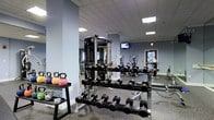 This fitness center is a thoughtfully designed workout facility built to accommodate a wide range of exercise needs. The space features a dropped ceiling with recessed panel lighting that provides bright, even illumination throughout. The walls are painted in a cool, neutral blue-grey tone, contributing to a focused and energizing atmosphere, while rubber flooring offers both durability and safety underfoot.

The strength training area is well-stocked with a full rack of dumbbells spanning a broad range of weights, complemented by kettlebells and medicine balls neatly organized on dedicated storage racks. A multi-functional cable machine serves as a centerpiece for resistance training, with large mirrors mounted along the wall to assist with form and to visually expand the space.

The cardio section is equipped with multiple machines, including ellipticals and upright exercise bikes, positioned to allow comfortable spacing between users. A wall-mounted flat-screen television is available for entertainment during workouts.

Natural light filters in through a window on the far wall, adding a welcoming element to the otherwise interior-focused layout. An emergency exit sign and clearly marked door ensure safe navigation throughout the facility. Overall, this fitness center offers a clean, organized, and fully functional environment suitable for a comprehensive training routine.