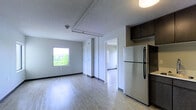 This single-floor unit presents a clean, functional layout designed for efficient everyday living. The main living area is anchored by light-toned hardwood flooring that runs throughout the space, reflecting natural light and contributing to an open, airy atmosphere. A square window overlooks a lush, green outdoor setting, bringing in ample daylight and providing pleasant views of the surrounding landscape.

The textured ceiling adds subtle character to the space, while exposed piping runs horizontally overhead, lending a modest industrial touch. Neutral grey walls keep the interior feeling bright and versatile. A smoke detector is visibly mounted on the ceiling, reflecting practical safety considerations built into the unit's design.

The kitchen area is positioned to one side and features dark espresso-toned upper cabinetry paired with a full-size stainless steel refrigerator, offering a clean contrast against the lighter surroundings. An adjacent doorway connects to a secondary room, which benefits from its own natural light source through a narrow sidelight window. A closet with a white door provides accessible storage. Black baseboards frame the flooring throughout, adding a subtle defining detail to the overall aesthetic.