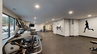 Located at 2988 Radio Rd in River Falls, Wisconsin, this fitness center offers a generously sized, single-floor exercise space designed to accommodate a full range of workout activities. The room features durable rubber flooring throughout, providing a safe and functional surface for all types of exercise. Recessed lighting illuminates the space evenly, complemented by large windows along one wall that invite natural light and provide outdoor views of the surrounding community. A row of treadmills and elliptical trainers lines one side of the room, while a multi-station cable and strength machine anchors the equipment zone. Additional amenities include a rowing machine, adjustable weight bench, dumbbells, and stability balls, supporting both cardio and resistance training needs. A wall-mounted flat-screen television is positioned for easy viewing from the cardio area. The open central floor space allows for stretching, mat work, or functional training. A full-length mirror along one wall enhances the sense of openness. Motivational quotes and a bold runner silhouette are featured as wall art, contributing an energizing ambiance. A water fountain is conveniently accessible within the space, and multiple doors provide easy entry and exit. The overall design is clean, modern, and purposefully laid out to serve a wide range of fitness goals.