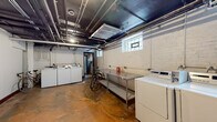This basement-level laundry facility offers a practical and utilitarian space designed to serve multiple users efficiently. The room is characterized by its industrial aesthetic, with an exposed dark-painted ceiling revealing an intricate network of pipes, ducts, and conduits running overhead. The concrete floor, with its warm amber and ochre tones, adds a raw, functional character to the space.

Multiple commercial-grade washing machines and dryers are arranged along the painted white brick walls, providing ample laundry capacity. A stainless steel utility table positioned centrally offers a dedicated surface for sorting and folding laundry. Small hopper-style windows along one wall allow modest natural light to filter into the space.

Additional features include a dedicated electrical panel mounted near the entry door, providing accessible utility access. A separate adjoining area visible through a doorway suggests additional storage or utility space. The room also accommodates bicycle storage, making it a versatile multi-purpose area. Dark-painted entry doors and brick accents contribute to the space's sturdy, no-frills character. Overall, this well-equipped basement facility is designed with practicality and shared-use functionality at its core.