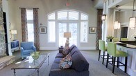 This single-story space makes a strong architectural statement with its dramatically tall ceilings and a striking arched window ensemble that serves as the room's focal point. The expansive window grouping, framed by colorful patterned drapery panels, allows abundant natural light to flood the interior while offering open views of the surrounding exterior. The light neutral wall tones complement the bright, airy atmosphere throughout.

The seating area is thoughtfully arranged around a plush, charcoal-gray sectional sofa accented with decorative throw pillows and a patterned blanket. A glass-topped coffee table with a delicate metal frame anchors the central gathering space, while a classic table lamp adds warmth to the arrangement. Additional seating options include a powder-blue accent chair and lime-green bar-height chairs, introducing vibrant pops of color against the soft, neutral backdrop.

Framed artwork adorns the walls on either side of the window feature, adding a curated, polished character to the space. The combination of high ceilings, generous natural light, eclectic furnishings, and inviting layout creates a versatile, welcoming environment well suited for relaxation and social interaction.