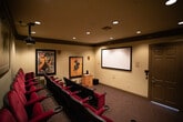 This private home theater is designed to replicate the experience of a classic cinema while offering the comfort and exclusivity of a personal screening room. The space is organized across two distinct zones: a lobby entry area and the main screening room, connected by a short set of steps.

The entry lobby sets a cinematic tone immediately, featuring dark wood crown molding, recessed lighting, and vintage-style framed posters decorating the walls — including classic French advertising prints. A popcorn machine adds an authentic movie-house touch, further enhancing the immersive atmosphere.

The main screening room continues the theater aesthetic with multiple rows of plush, tufted red-and-black theater-style seats arranged in a tiered, stadium configuration for unobstructed sightlines. Cupholders are integrated into each armrest. A ceiling-mounted projector delivers the picture onto a large, wall-mounted projection screen at the front of the room. Additional vintage cinema posters adorn the side walls, reinforcing the classic movie house ambiance.

The room is finished with dark patterned carpet, dark crown molding along the ceiling perimeter, and recessed lighting throughout. A small wooden media cabinet sits near the screen wall. The overall atmosphere is intimate, dramatic, and thoughtfully designed for an authentic cinematic experience.