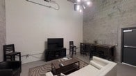 This single-floor space showcases a striking industrial aesthetic defined by its dramatically high ceilings and exposed concrete block walls, one painted white and the other left in its natural gray tone, adding textural contrast and visual depth. A modern sputnik-style chandelier hangs prominently from above, serving as a focal point and illuminating the open layout with a contemporary flair. The living area is anchored by a low-profile media console housing a flat-screen television, positioned against the white block wall. A rectangular wooden coffee table sits atop a patterned area rug, surrounded by white upholstered seating that provides a clean, modern counterpoint to the rugged surroundings. Adjacent to the living area, a tall wooden bar-height table paired with industrial-style metal stools creates a casual dining or workspace zone. Exposed conduit and piping trace along the upper walls, reinforcing the space's raw, urban character. The polished concrete flooring ties the elements together, contributing to the loft-like atmosphere. Overall, the space balances industrial architectural features with carefully chosen furnishings to create a functional and visually engaging environment.