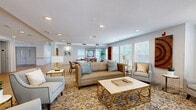 Located at 7401 E 26th Ave in Denver's Stapleton neighborhood, this well-appointed community lounge offers a warm and inviting atmosphere designed for both relaxation and social gatherings. The open, single-floor layout is anchored by a richly patterned area rug that defines the primary lounge area, where a tufted caramel-toned sofa, elegant armchairs, and a gold-accented coffee table create a refined yet comfortable seating arrangement. A wall-mounted flat-screen TV with a rustic media console serves as a focal point on one end of the room, accompanied by lush tropical plants that add a vibrant, natural touch throughout the space. Recessed lighting and multiple white-framed windows with blinds allow generous natural light to fill the room, enhancing its airy, open feel. The dining area features multiple wood tables with upholstered chairs, ideal for shared meals or group activities, and is further accented by framed wall art and decorative wooden pieces. A dedicated entryway with smooth hardwood flooring leads to elevator access and exits, offering seamless connectivity. The overall ambiance is elegant and welcoming, blending neutral tones, gold accents, and thoughtful décor to create a sophisticated communal space.