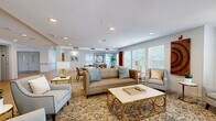 Located at 7401 E 26th Ave in Denver's Stapleton neighborhood, this well-appointed community lounge offers a warm and inviting atmosphere designed for both relaxation and social gatherings. The open, single-floor layout is anchored by a richly patterned area rug that defines the primary lounge area, where a tufted caramel-toned sofa, elegant armchairs, and a gold-accented coffee table create a refined yet comfortable seating arrangement. A wall-mounted flat-screen TV with a rustic media console serves as a focal point on one end of the room, accompanied by lush tropical plants that add a vibrant, natural touch throughout the space. Recessed lighting and multiple white-framed windows with blinds allow generous natural light to fill the room, enhancing its airy, open feel. The dining area features multiple wood tables with upholstered chairs, ideal for shared meals or group activities, and is further accented by framed wall art and decorative wooden pieces. A dedicated entryway with smooth hardwood flooring leads to elevator access and exits, offering seamless connectivity. The overall ambiance is elegant and welcoming, blending neutral tones, gold accents, and thoughtful décor to create a sophisticated communal space.