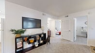 This single-floor residence offers a thoughtfully designed layout with bright, white walls and soft carpeting throughout most of the living spaces. The open living and dining area serves as the heart of the home, featuring a ceiling fan, a wall-mounted flat-screen TV above a storage console, and a compact dining set positioned near the entry — creating a welcoming, multifunctional gathering space. Adjacent to this area, a comfortable sitting room provides a relaxed retreat with a large sectional sofa, a patterned area rug, and a generous window that brings in natural light and views of surrounding greenery. The kitchen is clean and efficient, outfitted with white cabinetry, a white electric range, and wood-look flooring that adds warmth to the space. The primary bedroom is a serene retreat, complete with a ceiling fan, a large framed mirror, dual windows, and plush carpeting. A secondary bedroom doubles as a functional workspace, featuring a built-in desk, shelving, a ceiling fan, and a bright window overlooking outdoor greenery. The full bathroom is well-appointed with a white vanity, a tub/shower combination, and wood-look flooring. Throughout, the residence maintains a cohesive, light-filled aesthetic with a clean and modern sensibility.