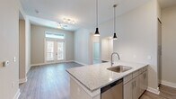 Located at 3915 William E Murray Blvd in Charleston, SC, this well-appointed 2-bedroom, 2-bathroom residence offers a thoughtfully designed single-floor layout with modern finishes throughout. The open-concept kitchen and living area serves as the heart of the home, anchored by a large granite-topped island with a stainless steel sink and stylish pendant lighting. The kitchen is fully equipped with stainless steel appliances, including a refrigerator, range, microwave, and dishwasher, complemented by contemporary cabinetry and durable wood-style flooring. The living area features soaring ceilings, a ceiling fan, and French doors with transom windows that flood the space with natural light and provide access to an outdoor balcony. The bedrooms are carpeted for comfort, with the primary bedroom offering generous proportions, tall ceilings, a walk-in closet, and a private en-suite bathroom featuring a dual-sink granite vanity, wood-style flooring, and a glass-enclosed shower. The second bathroom includes a full tub-shower combination and a granite vanity. Built-in shelving in a dedicated nook adds practical storage. Neutral tones and clean architectural lines create a bright, cohesive ambiance throughout.